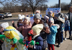 Dzieci maszerują ulicami miasta w przemarszu wiosennym, dwie dziewczynki trzyma w ręku kukłę Pani Wiosny oraz maskotkę Krasnala Hałabały.